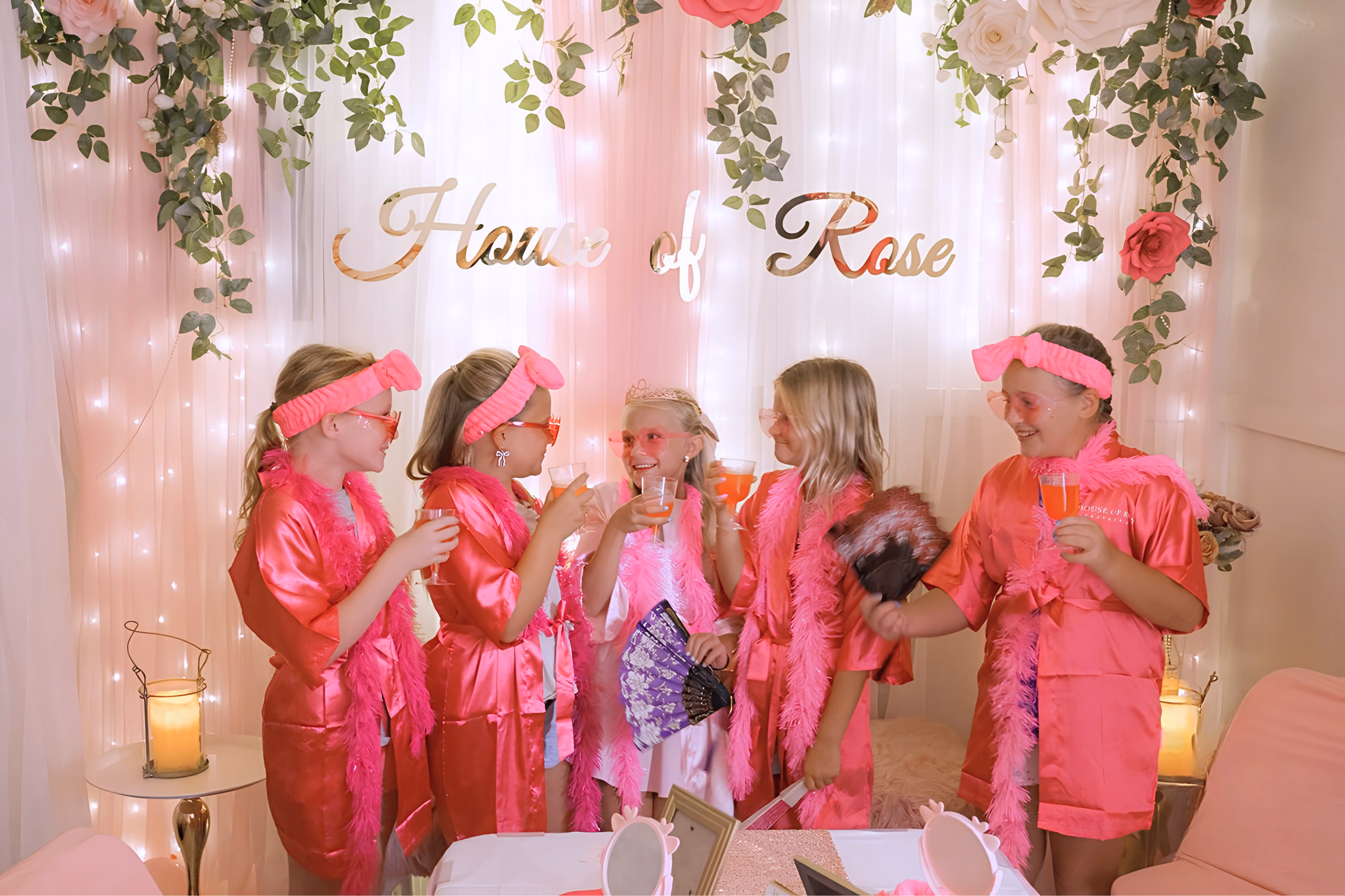Five children in pink satin robes laughing together during a sparkling pink lemonade toast in front of the House of Rose backdrop.