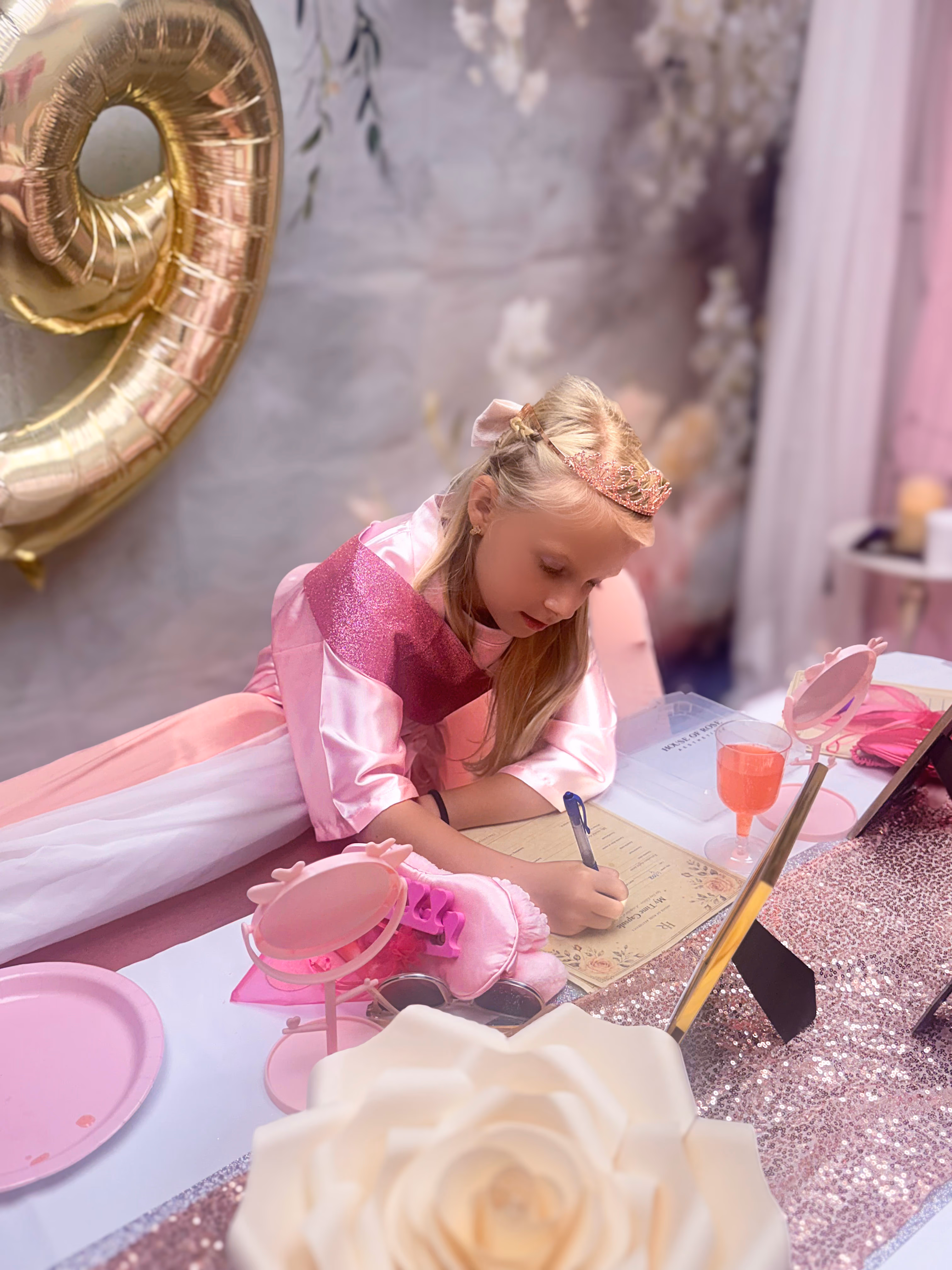 A child in a pink event robe writing a time-capsule note at the decorated Little Roses table.